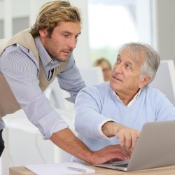 Ein älterer Herr sitzt vor einem Laptop. Er schaut fragend zu einem jungen Mann, der neben ihm auf den Bildschirm schaut.