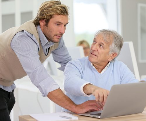 Schulungssituation. Ein älterer Herr sitzt vor einem Laptop. Er schaut fragend zu einem jungen Mann auf, der neben ihm steht und sich unterstützend zu ihm herunterbeugt.