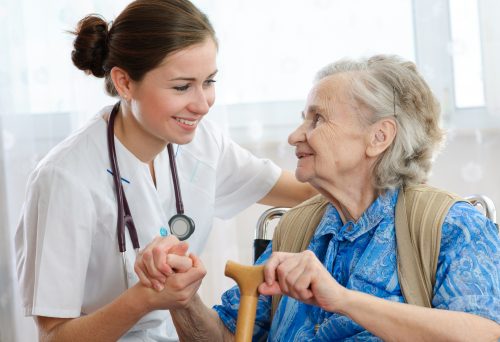 Eine Pflegerin mit Stethoskop hält die Hand einer alten Dame in einem Stuhl. Die Frauen lächeln sich an.