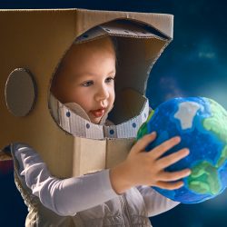 Ein Kleinkind hat einen aus Karton gebastelten Raumfahrer-Helm auf dem Kopf. In seinen Händen hält es das Modell einer Erdkugel.