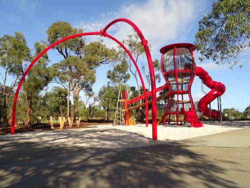 Ein Spielplatz mit Schaukel, Hängebrücke, Kletterturm, einer gewundenen Tunnelrutsche und einer Kleinkind-Rutsche. Die Spielgeräte bestehen aus rotem Metall. Hinter dem Spielplatz stehen Bäume.