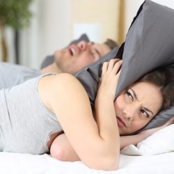Eine Frau liegt im Bett und hält sich genervt mit einem Kopfkissen die Ohren zu. Neben ihr liegt ein Mann auf dem Rücken und schnarcht.