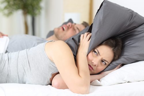 Eine Frau liegt im Bett und hält sich genervt mit einem Kopfkissen die Ohren zu. Neben ihr liegt ein Mann auf dem Rücken und schnarcht.