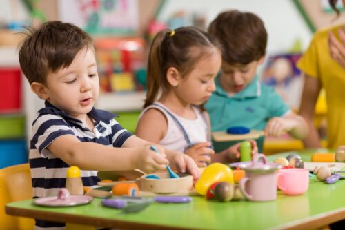 Zwei Jungen und ein Mädchen im Kindergartenalter spielen an einem grünen Tisch. Auf dem Tisch liegen viele bunte Utensilien für eine Spielküche.