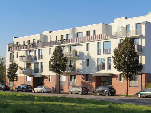 Ein modernes Wohngebäude mit zwei Eingängen und vier Etagen, Balkons und ein Flachdach mit Terrassen. Vor dem Gebäude stehen Autos zwischen jungen Bäumen.