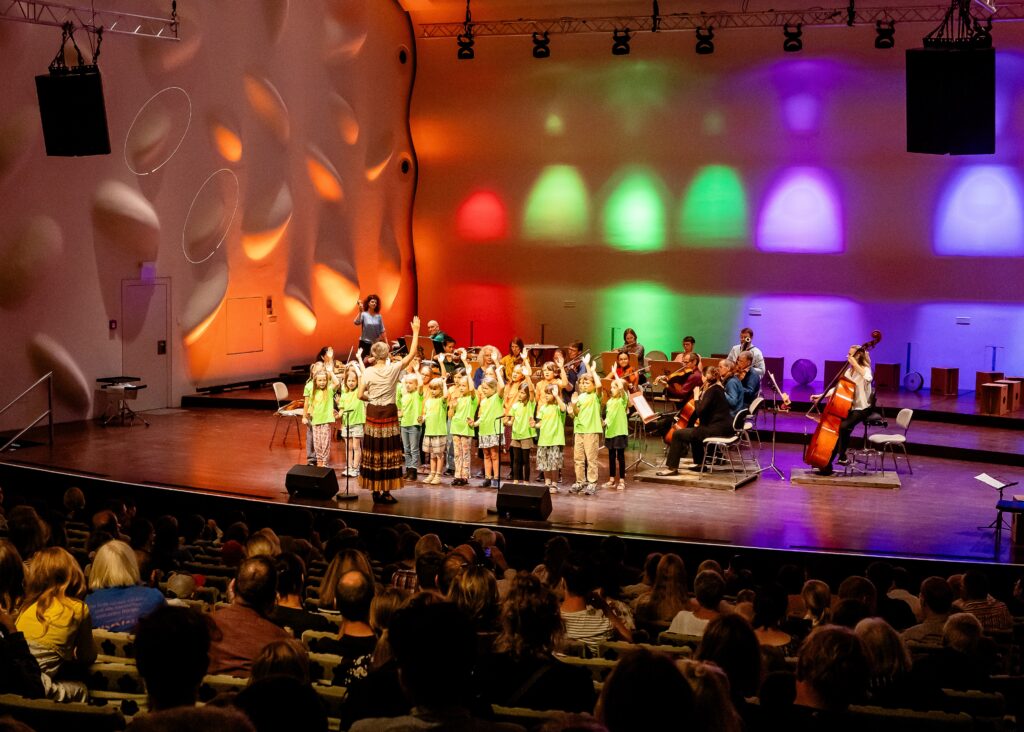 Ein Kinderchor singt auf einer Bühne, dahinter musizieren erwachsene Musiker*innen.