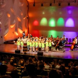 Ein Kinderchor singt auf einer Bühne, dahinter musizieren erwachsene Musiker*innen.