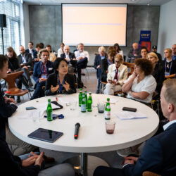 Sechs Menschen sitzen an einem runden Tisch in einem Gespräch, im Hintergrund hört eine Gruppe Menschen zu.